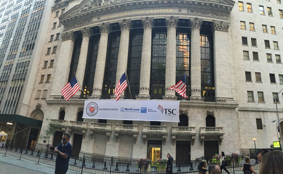 A banner across the facade of the New York Stock Exchange bearing the logos of Arms Wide Open Childhood Cancer Foundation, Guggenheim, Merrill Lynch, Goldman Sachs Asset Management, and TSC — TS Capital, LLC.